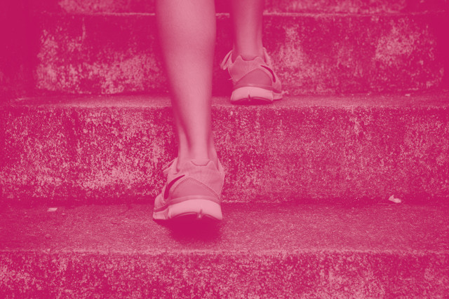 Ein rötlich eingefärbtes Foto von einer Person mit sportlichen Schuhen, die eine Treppe hinauf steigt. Die Beine sind zu sehen, da die Person eine kurze Hose trägt.