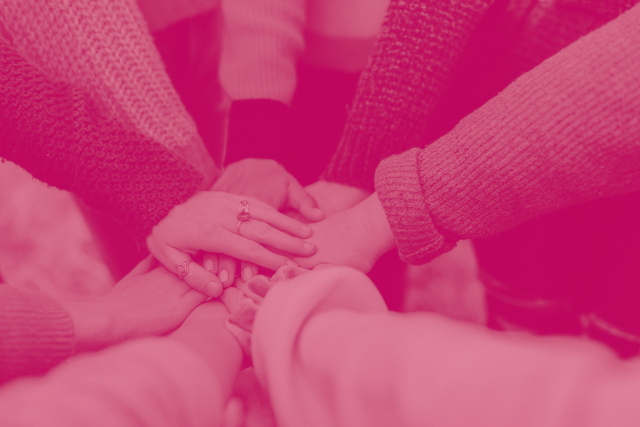Ein rötlich eingefärbtes Foto von mehreren Händen von Menschen die im Kreis zueinander stehen und die Hände in der Mitte aufeinander legen